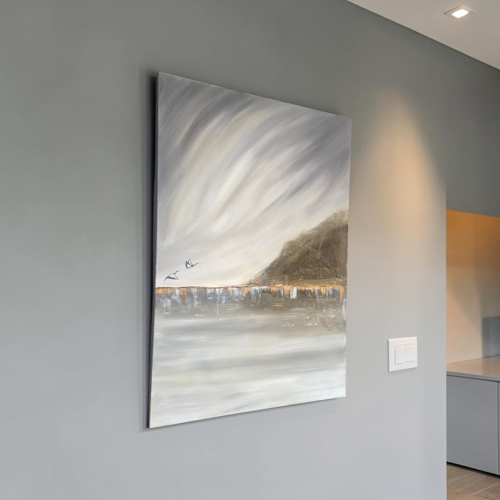 A vertical canvas painting of a moody, abstract landscape with swirling gray and white clouds, a dark hillside, and a reflective water line with small birds, displayed on a gray wall in a modern interior.