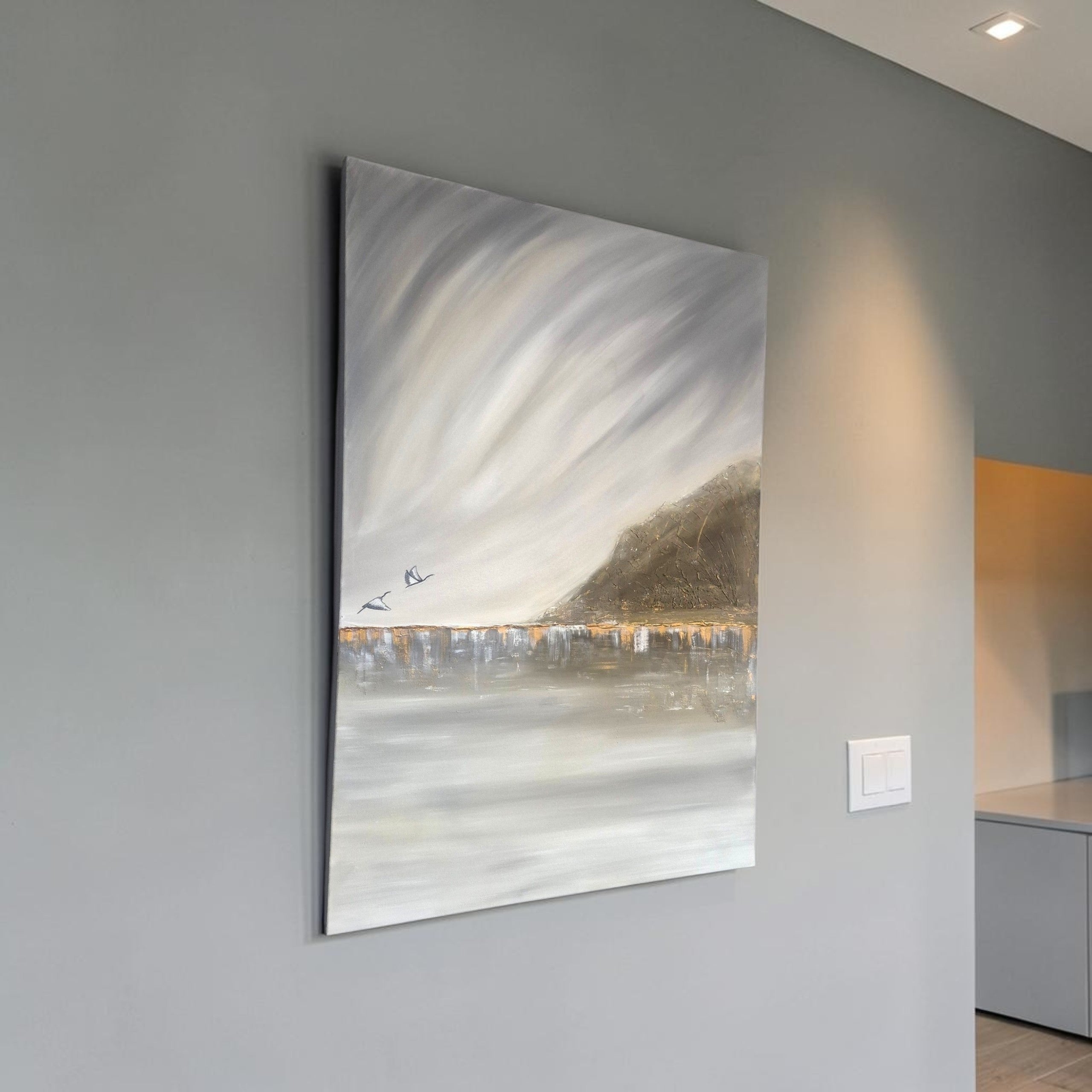 A vertical canvas painting of a moody, abstract landscape with swirling gray and white clouds, a dark hillside, and a reflective water line with small birds, displayed on a gray wall in a modern interior.