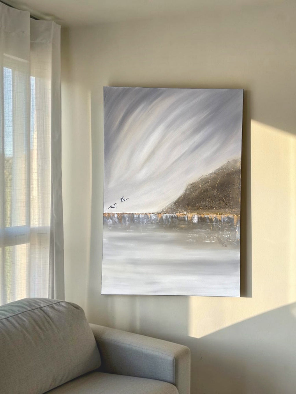 A vertical canvas painting of an abstract landscape with swirling gray and white clouds, a brown hill, and a reflective water line, displayed on a beige wall in a modern living room next to a window and sofa.