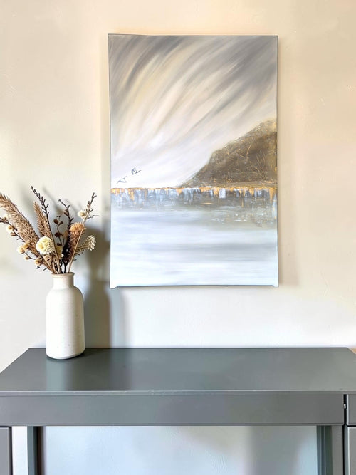 A vertical canvas painting of a coastal landscape with a gray sky, white water, and a brown hillside, displayed on a beige wall above a gray console table with a white vase of dried flowers.