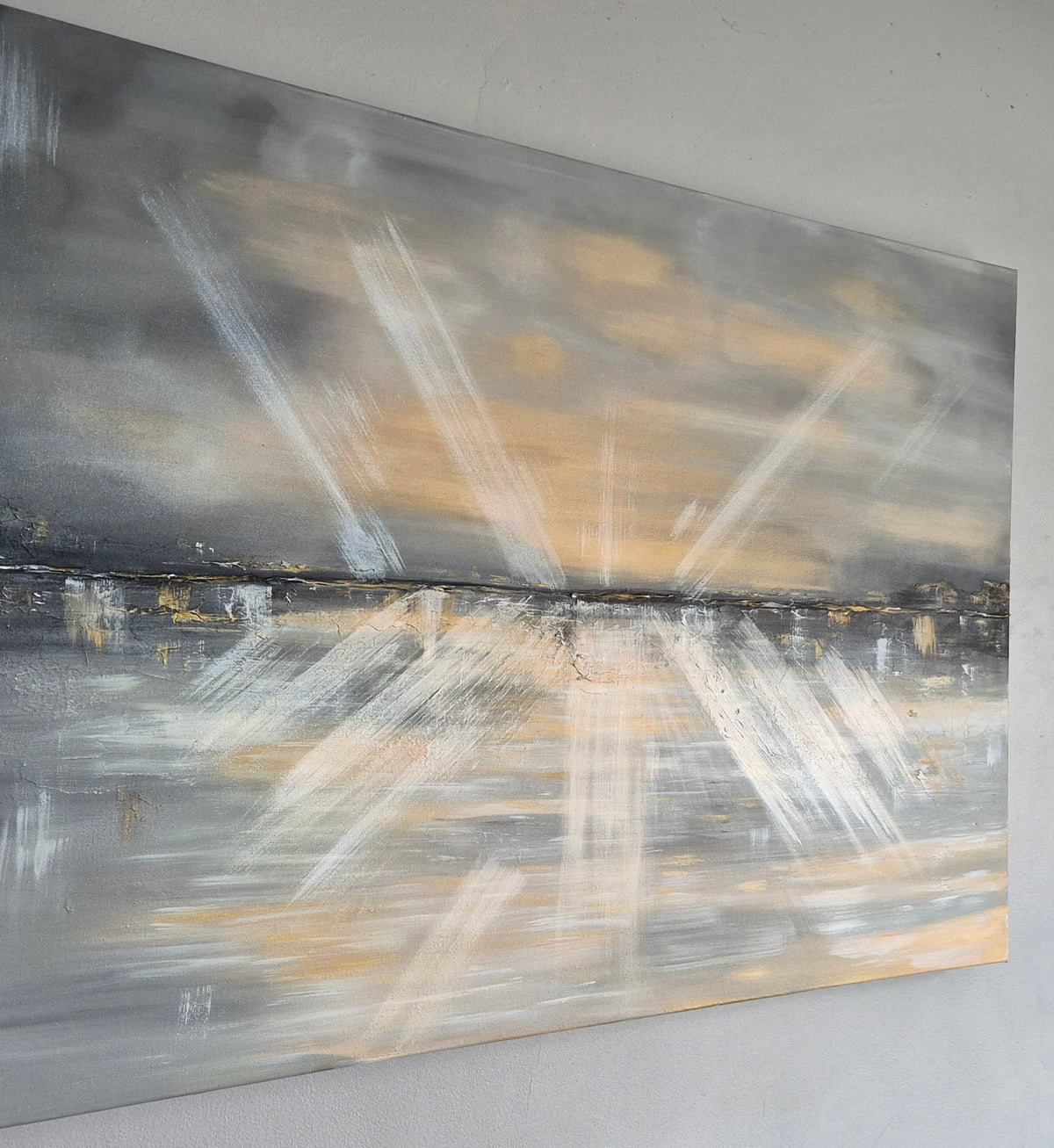 An abstract landscape painting on canvas, featuring radiating white brushstrokes over a gray and black background with warm beige and gold tones near the horizon, displayed on a light gray wall.