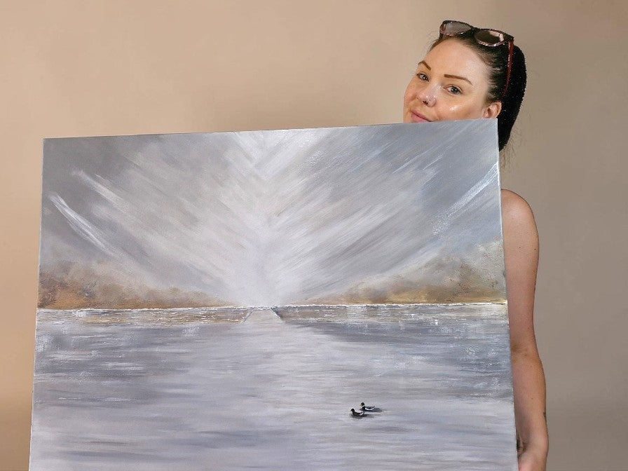 A woman holding up a large canvas painting of a calm lake or river scene with a bright sunburst over water and small boats near the bottom. The woman is partially visible behind the painting, wearing sunglasses on her head against a plain beige wall.