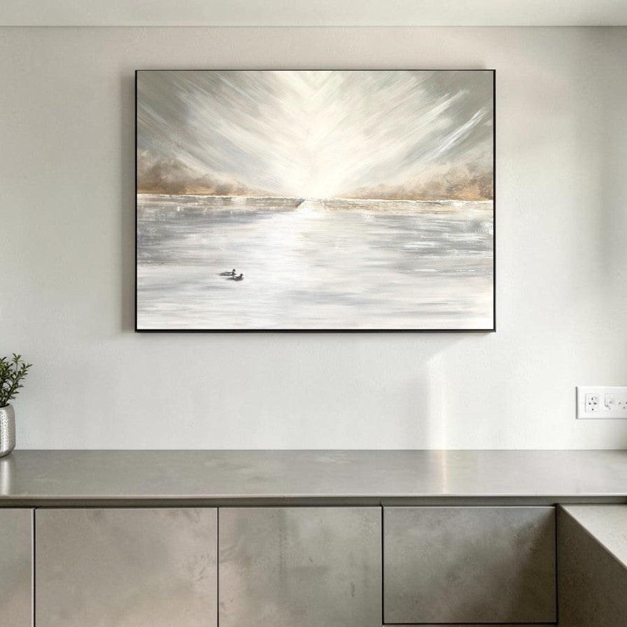 A framed abstract landscape painting hanging on a light gray wall above a metallic gray console table. The artwork depicts a bright sunlit water scene with soft gray, white, beige, and brown tones.
