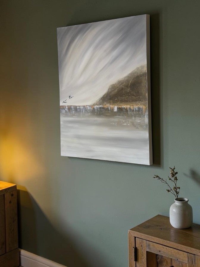 A vertical canvas wall art print depicting a misty coastal landscape with a dark hill and pale sky, hanging on a muted green wall above a wooden cabinet with a small vase.