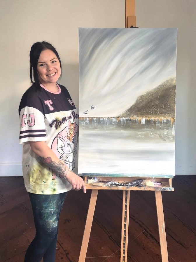 A woman stands beside a canvas painting on an easel in a studio, wearing a graphic t-shirt and dark pants. The painting depicts a misty landscape with a mountain and reflective water.