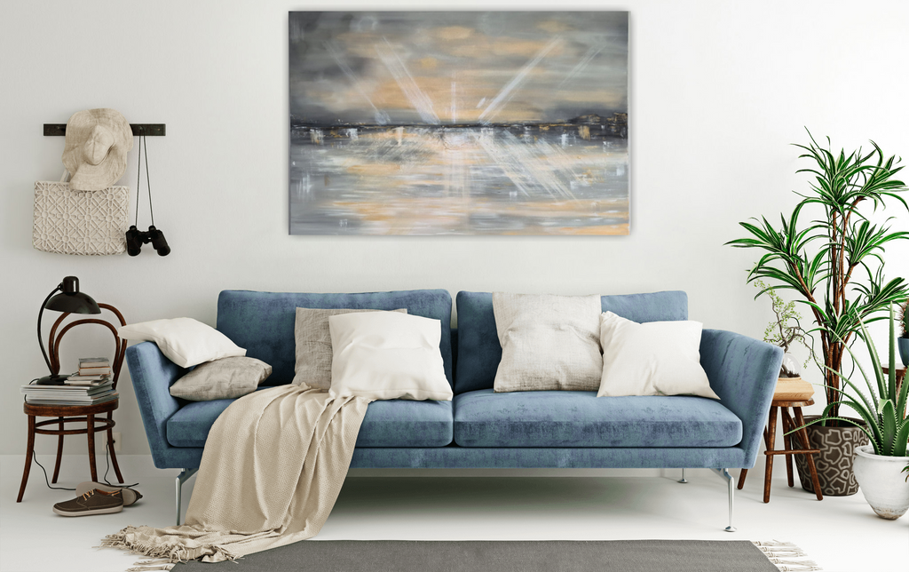 A blue fabric sofa in a bright, modern living room setting, styled with cream and gray throw pillows, a beige throw blanket, a small wooden side table with a lamp, and a tall potted plant on the right. A large abstract canvas print hangs on the white wall above the sofa.